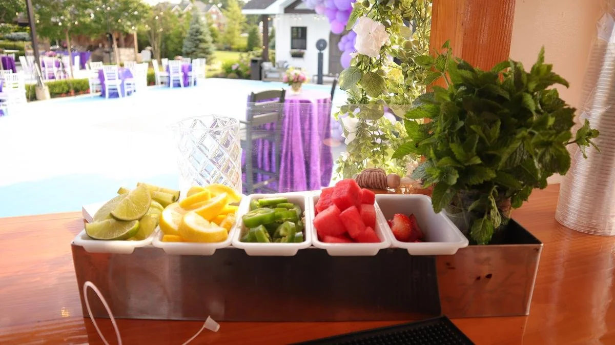 Tray of sliced citrus fruits, green beans, watermelon, and strawberries, along with a houseplant and a glass of water, in an outdoor dining area.