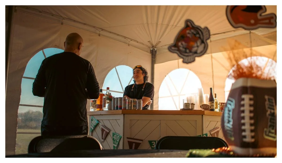 A man and a woman are standing and talking at a bar inside a tent decorated for a football game, with football-themed banners and decorations visible, and a football shaped drink container in the foreground.