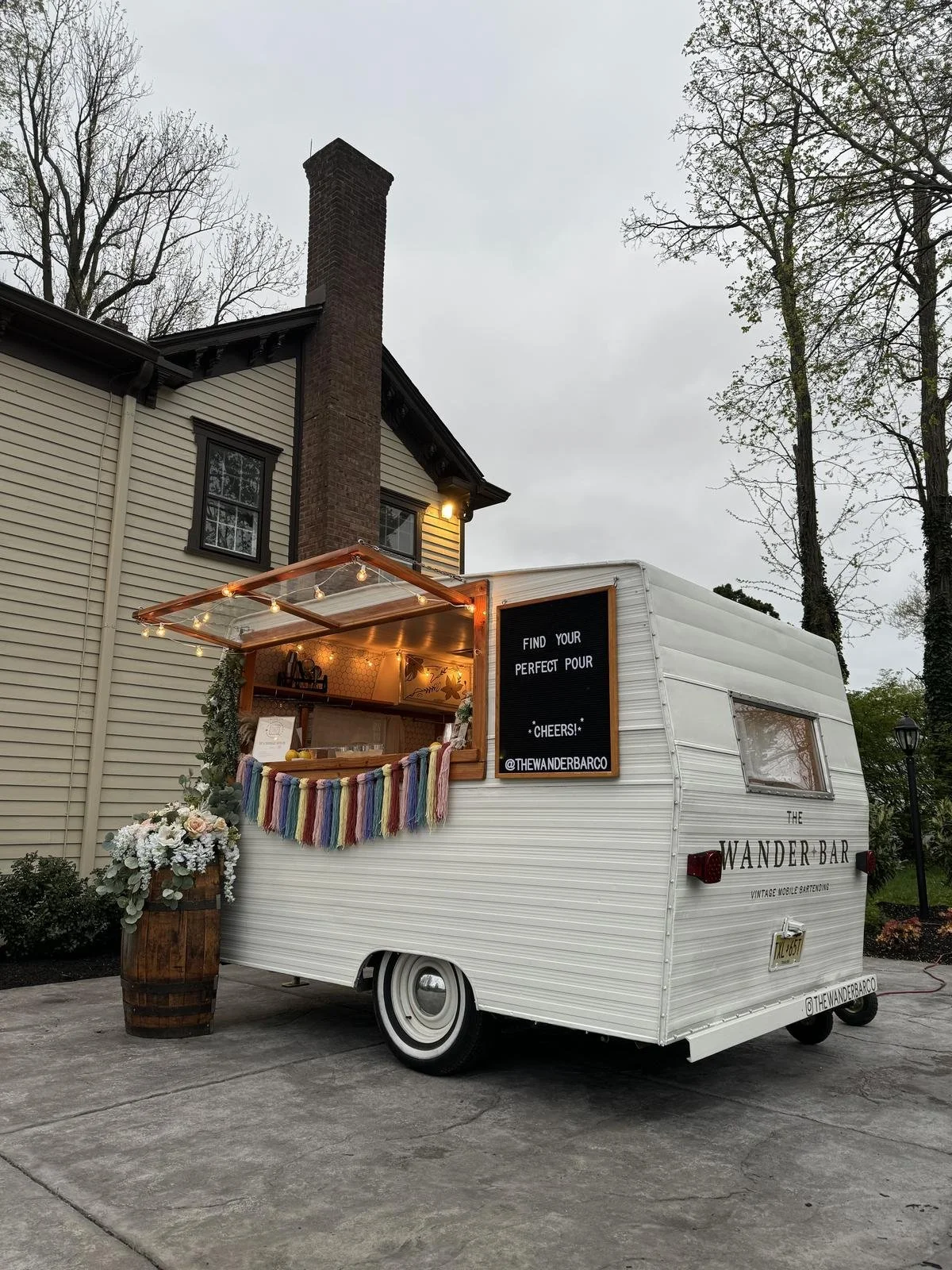 Vintage white mobile bar trailer labeled 'The Wander Bar' with a black sign on it that reads 'Find your perfect pour. Cheers! @THEWANDERBARCO'. The trailer has an open window with string lights and a rainbow-colored yarn decoration. There is also a w