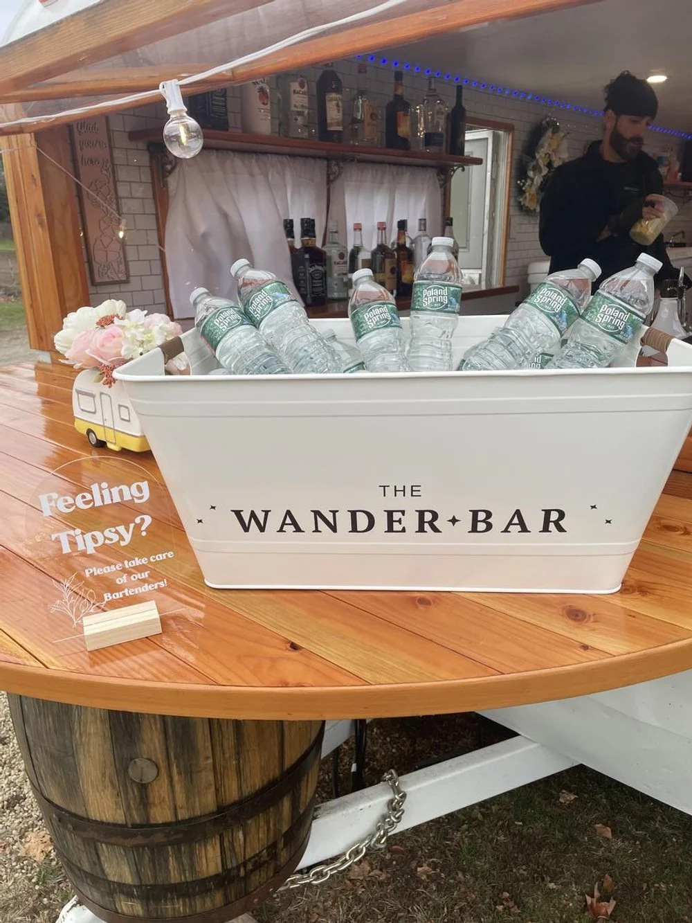 A white container labeled "The Wander Bar" filled with bottled water labeled "Poland Spring" on a wooden table at an outdoor bar or event setup. A small sign asking "Feeling tipsy? Please take care of our bartenders!" is also on the table. In the bac