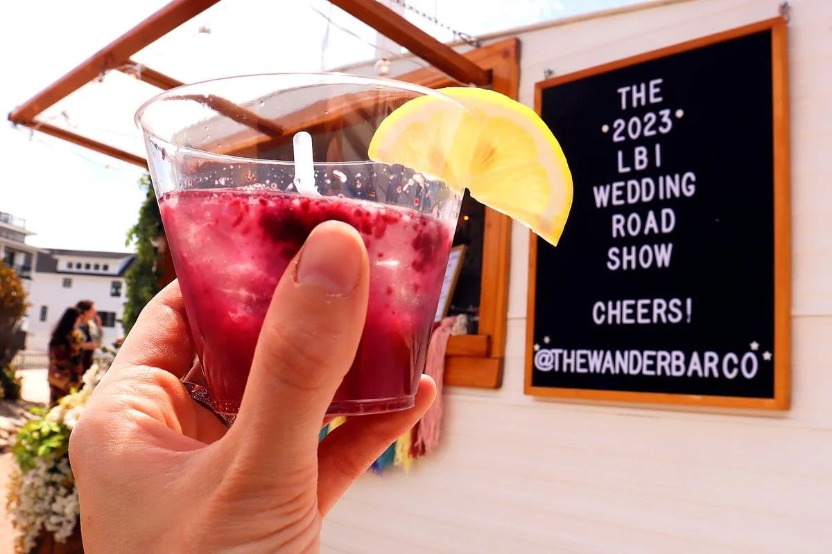 A person holding a pink drink in a clear plastic cup with a lemon wedge garnish, with a signboard in the background that reads 'The 2023 LBI Wedding Road Show Cheers! @THEWANDERBARCO'.