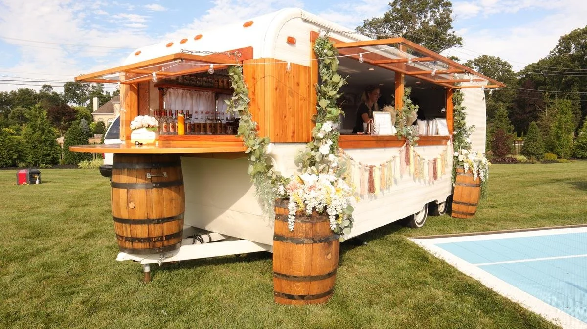 A decorated mobile bar trailer with wood accents and floral decorations, set up outdoors on a grassy area next to a swimming pool.
