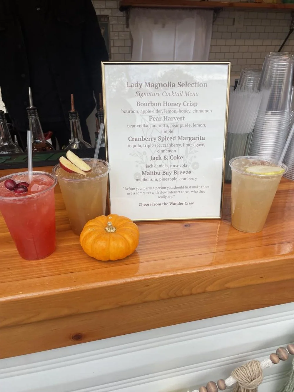 Three cocktails on a wooden bar with a pumpkin, a drink with apple slices, and a yellowish drink with lemon, behind a menu and plastic cups.