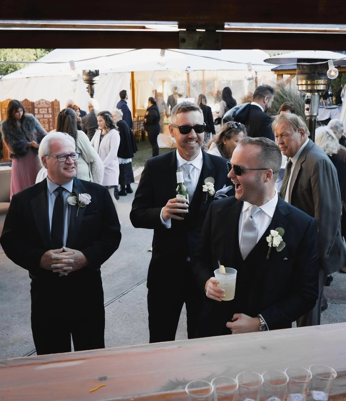 Group of men wearing suits and sunglasses, holding drinks and laughing at a wedding reception outdoors, with other guests in the background under a tent.