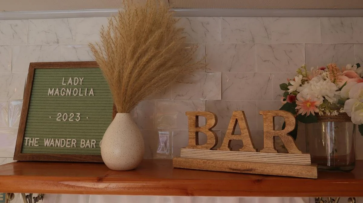 Decorative display on a wooden shelf with a framed letter board, a vase containing dried pampas grass, wooden letters spelling "BAR," and a flower arrangement in a metallic container.