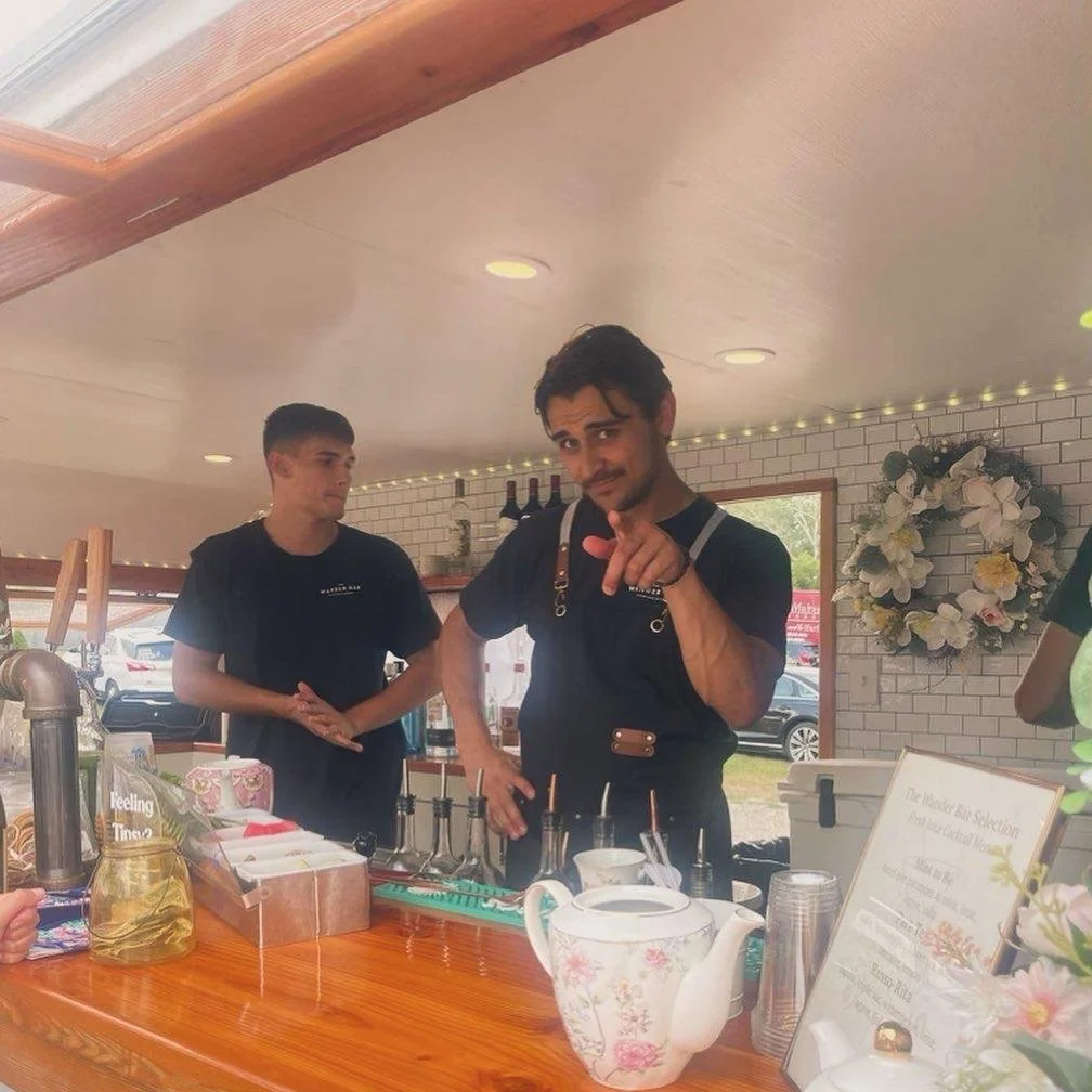 Two young men working at a coffee or tea stand inside a shop, with one man pointing at the camera. The stand has a teapot, cups, and syrups on the counter, with decorative flowers and a wreath on the wall in the background.