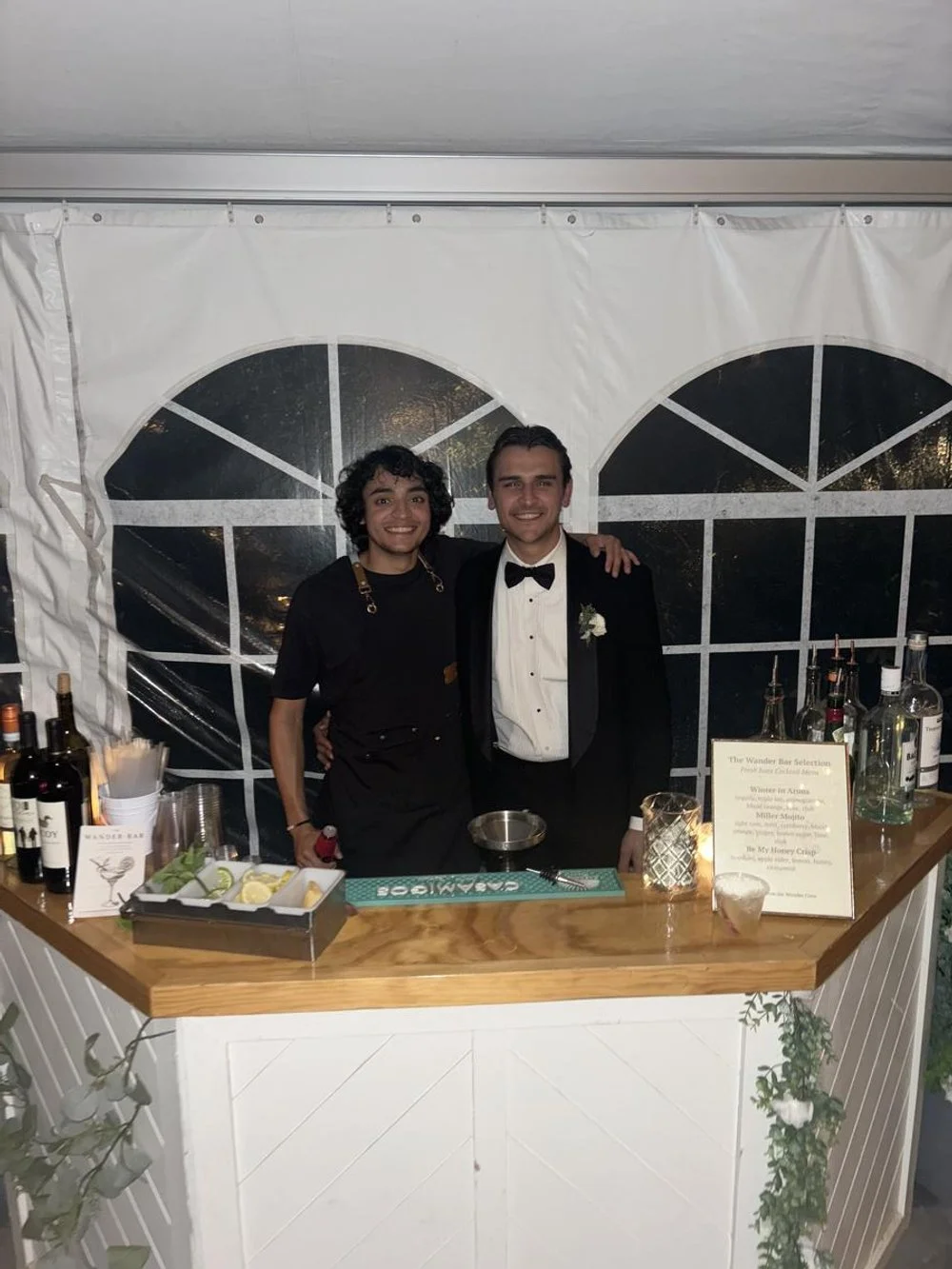 Two men standing behind a bar at a wedding reception, one dressed in a tuxedo and the other in a black shirt and apron, smiling and posing for a photo.