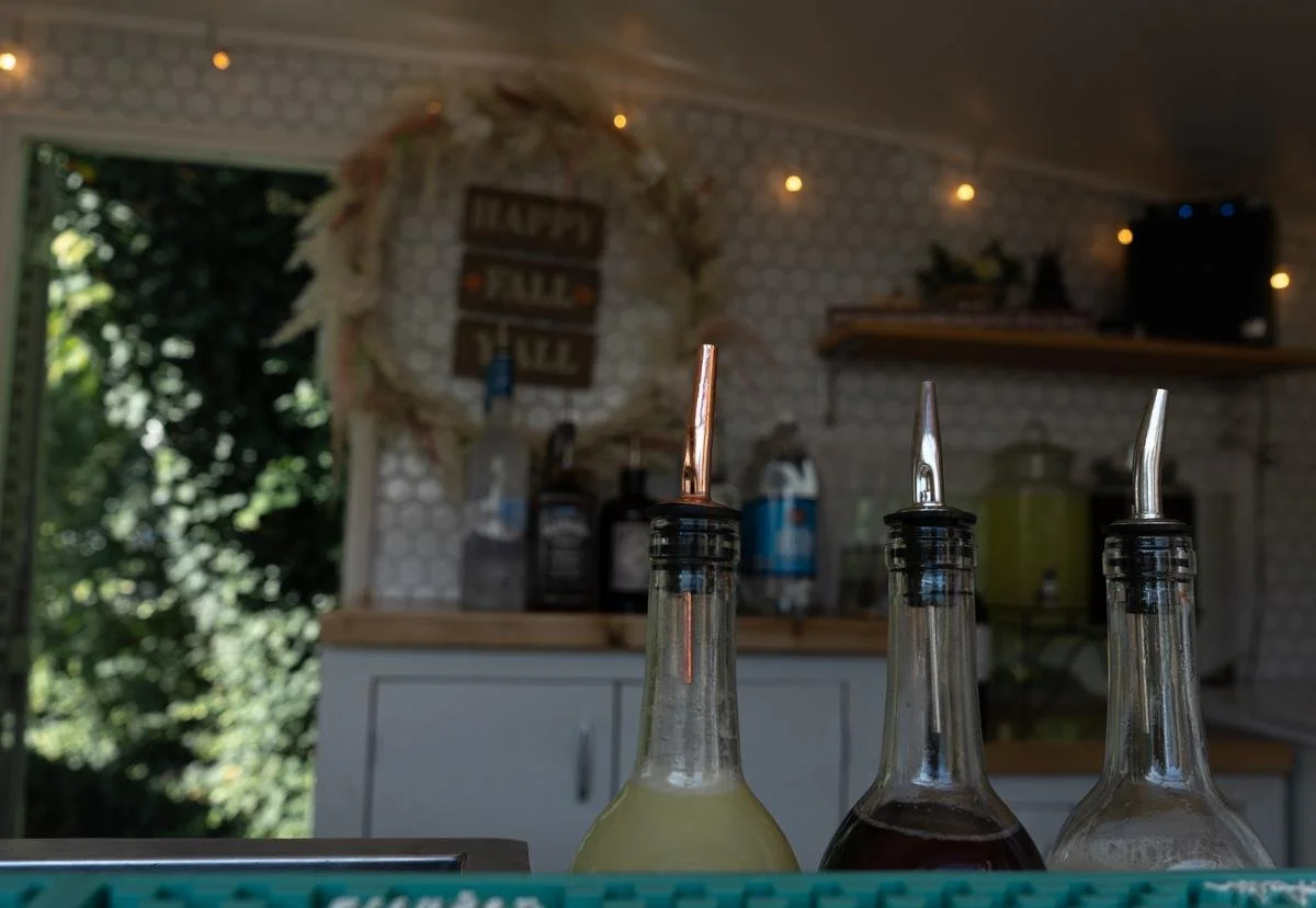 Three glass bottles with metal pour spouts contain different liquids, set on a counter in a cozy, decorated room with a window, plants, and wall hangings.