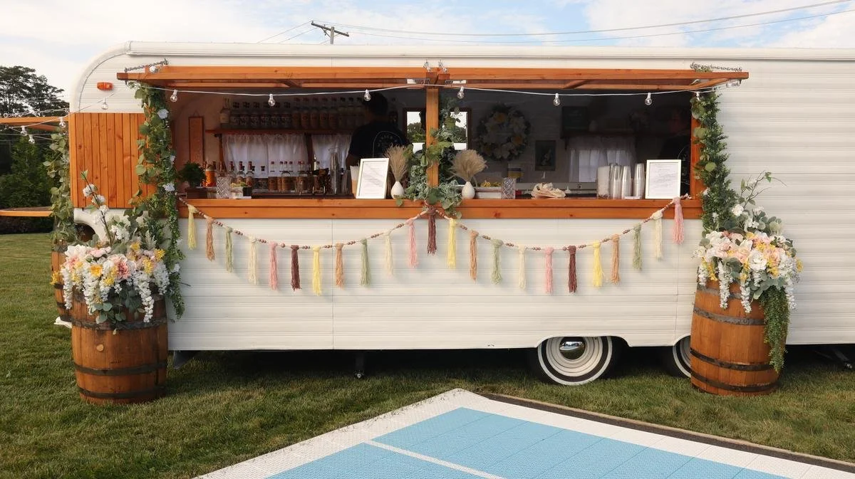 A decorated mobile food or drink stand set up outdoors on grass with floral arrangements on wooden barrels, string lights, and pastel-colored tassel garland.