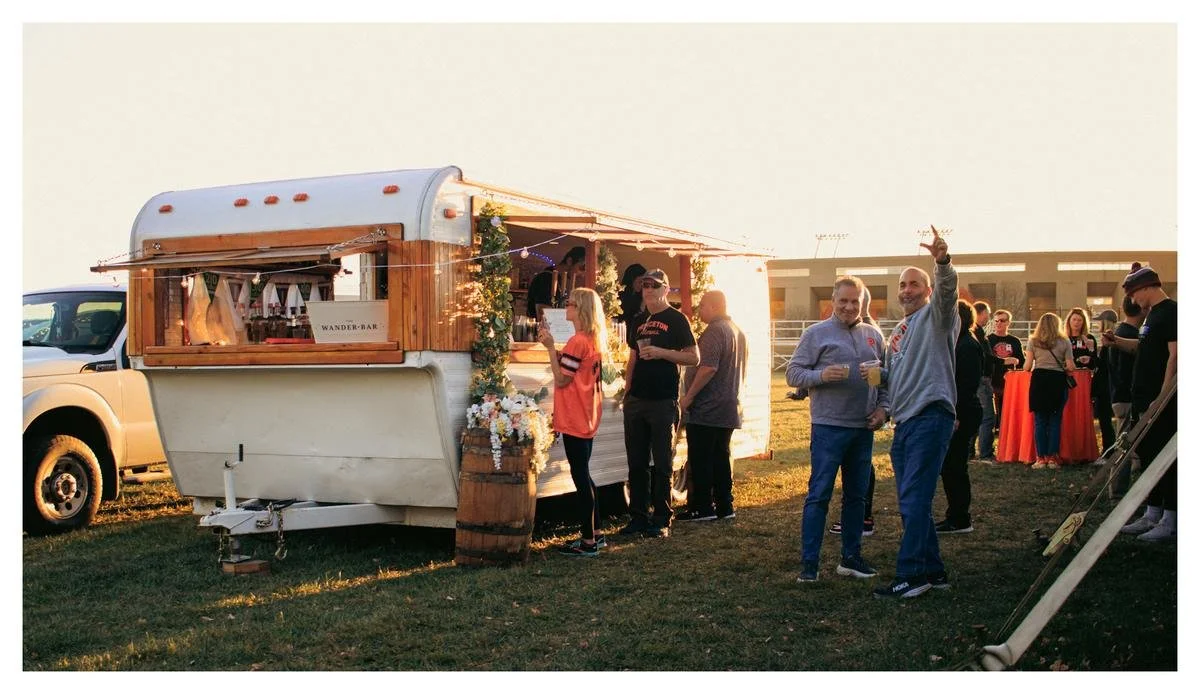 People gather around a wooden food truck at an outdoor event during sunset, some holding drinks and chatting.