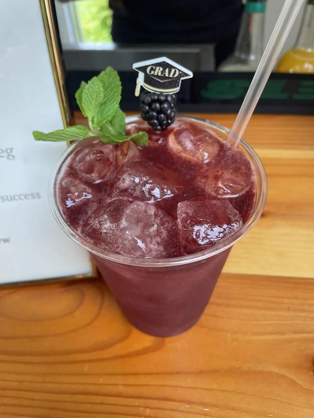 A purple berry-themed iced drink garnished with a mint sprig and a decorative black grape fedora with a 'GRAD' cap, served with a straw on a wooden table.