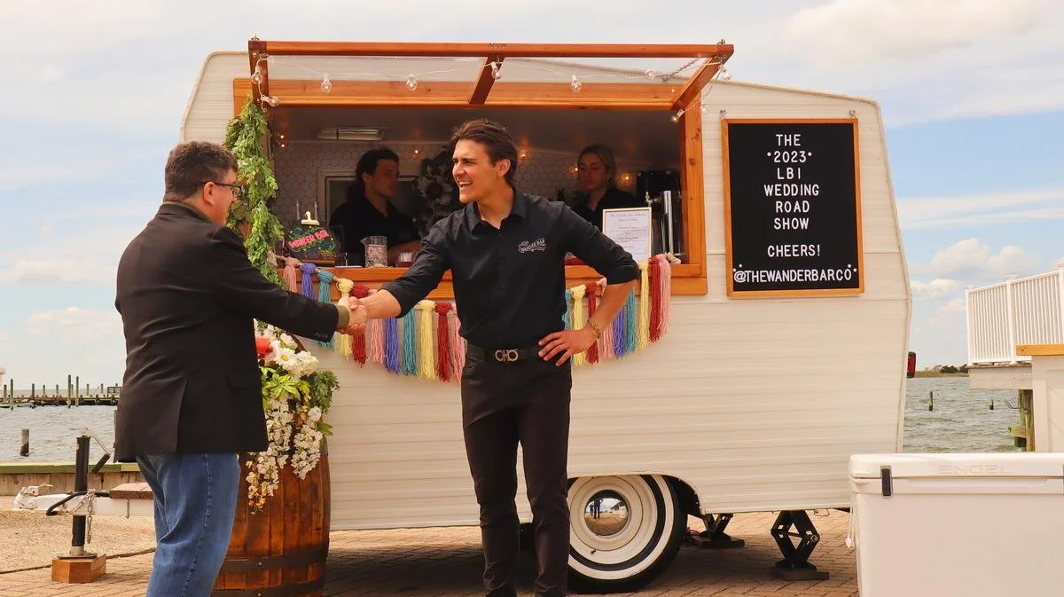 A man in a suit shaking hands with a man in a black shirt in front of a trailer with a sign that says 'The 2023 LBI Wedding Road Show'. Two women are inside the trailer, and there are colorful decorations and string lights hanging from the trailer.