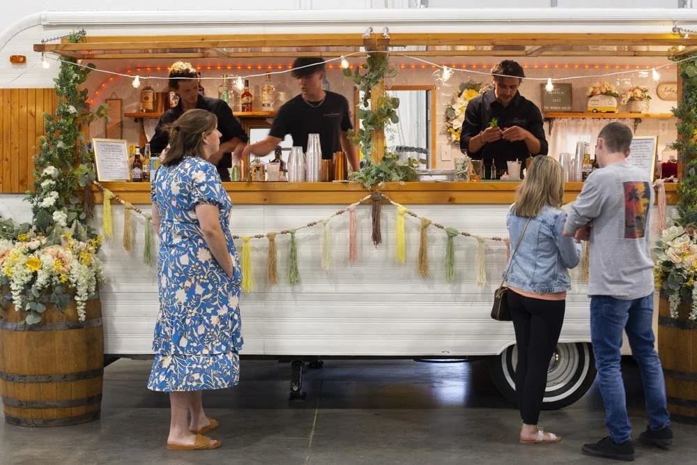 People ordering drinks at a decorated food truck bar with floral arrangements and string lights.