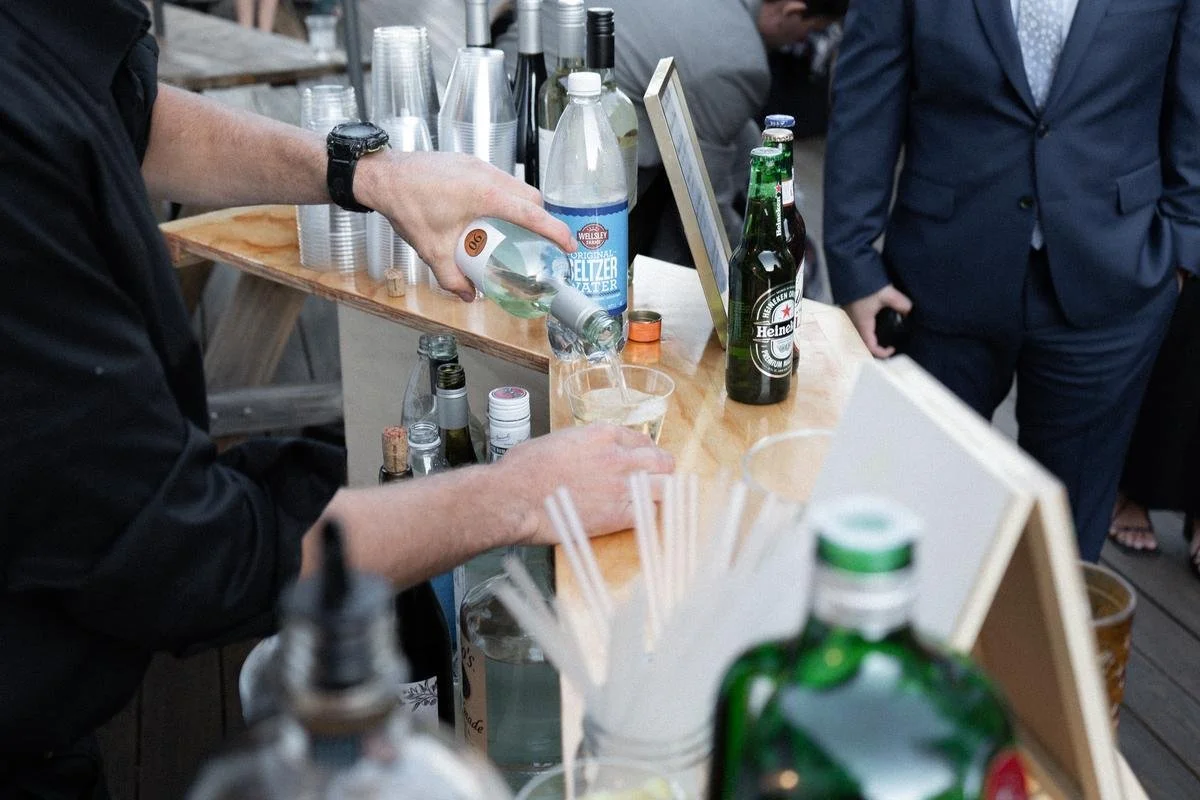 Person pouring a drink at a bar with bottles of alcohol and beer bottles on the counter, while other people stand nearby.