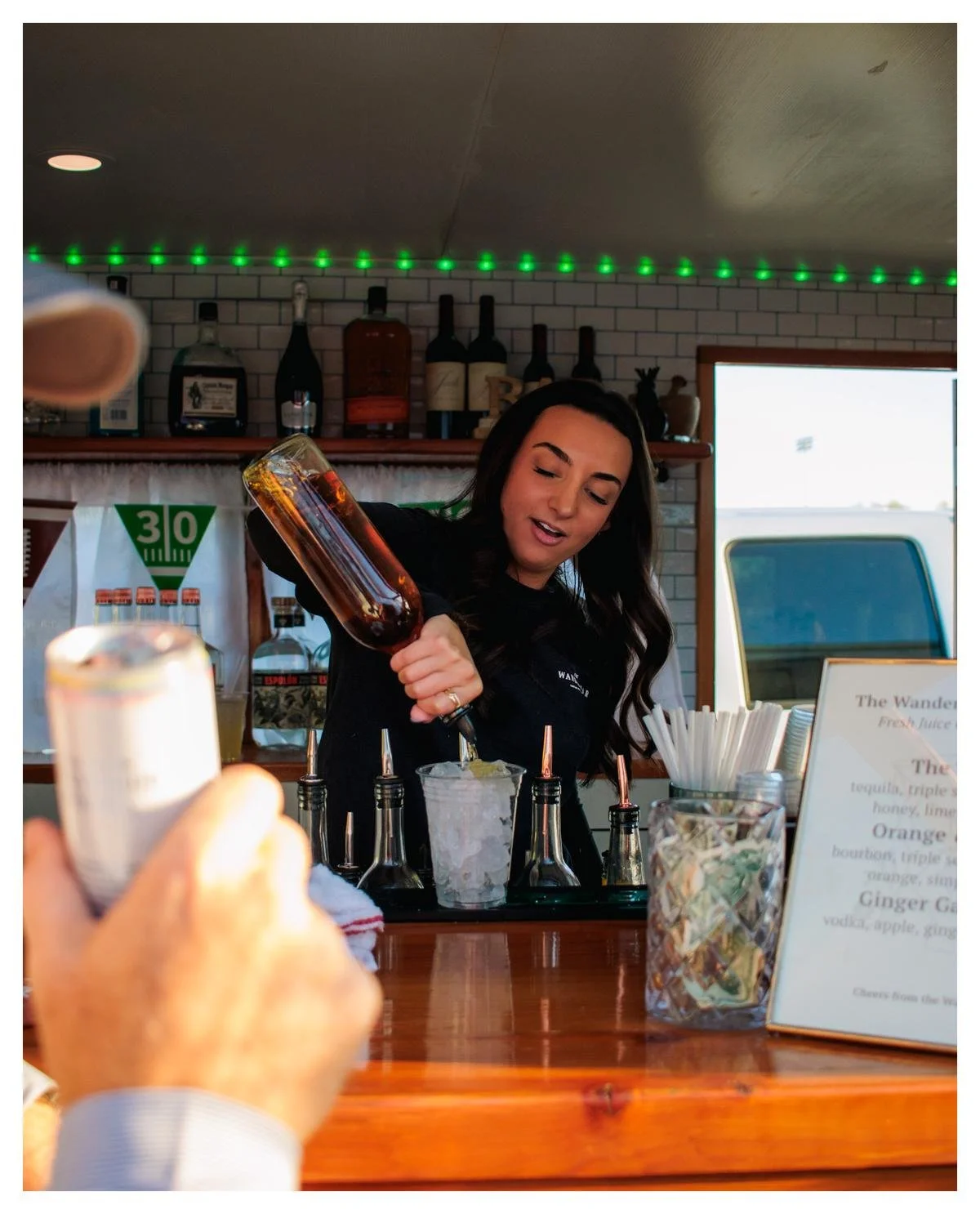 A woman behind a bar preparing a cocktail, pouring a dark liquid into a glass filled with ice. The bar has bottles of alcohol on the shelf, bar tools, and a menu visible.