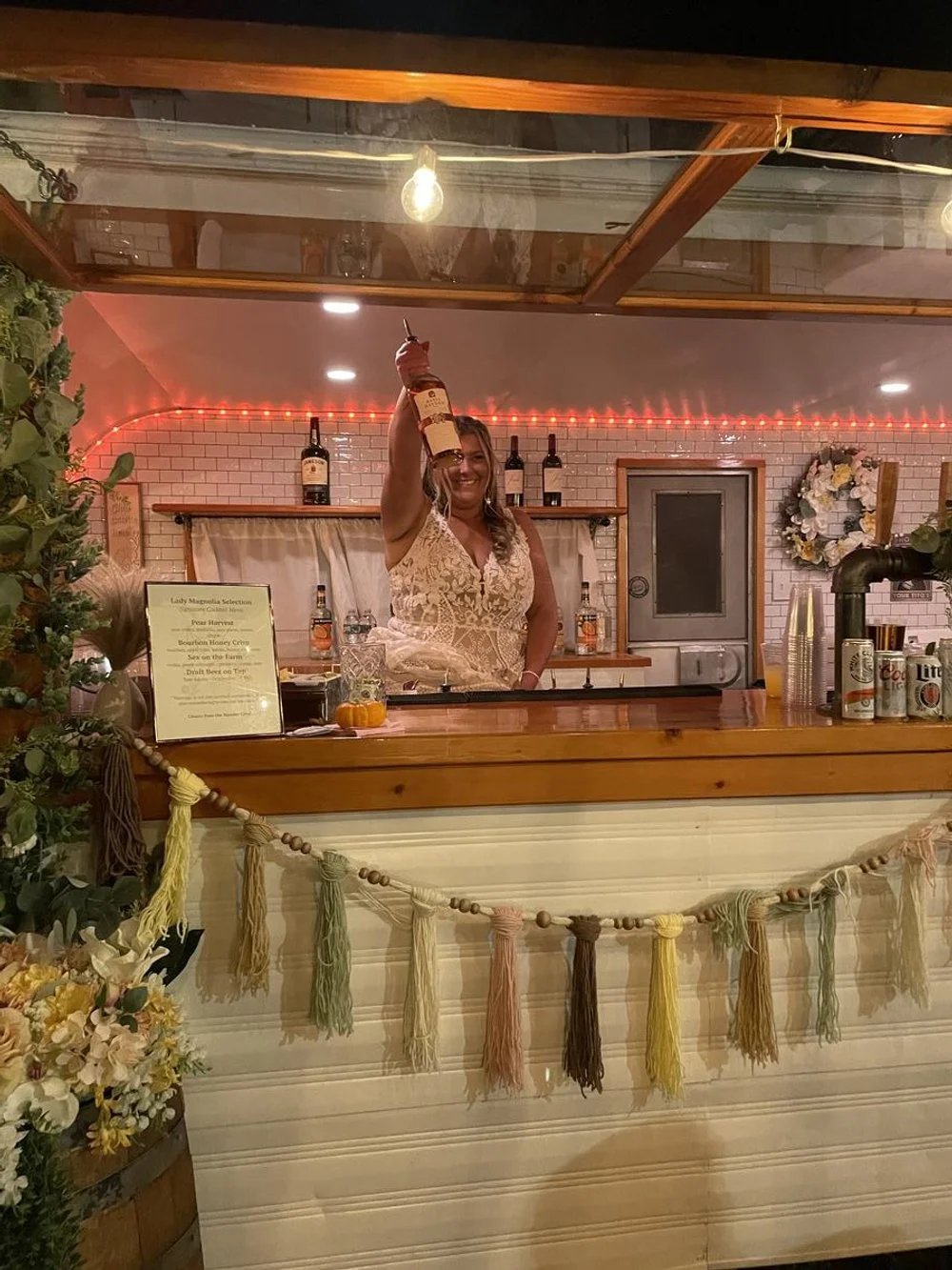 A woman in a lace dress raising a bottle behind a bar decorated with tassels and flowers, with bottles and glasses on the counter and a wreath on the wall.