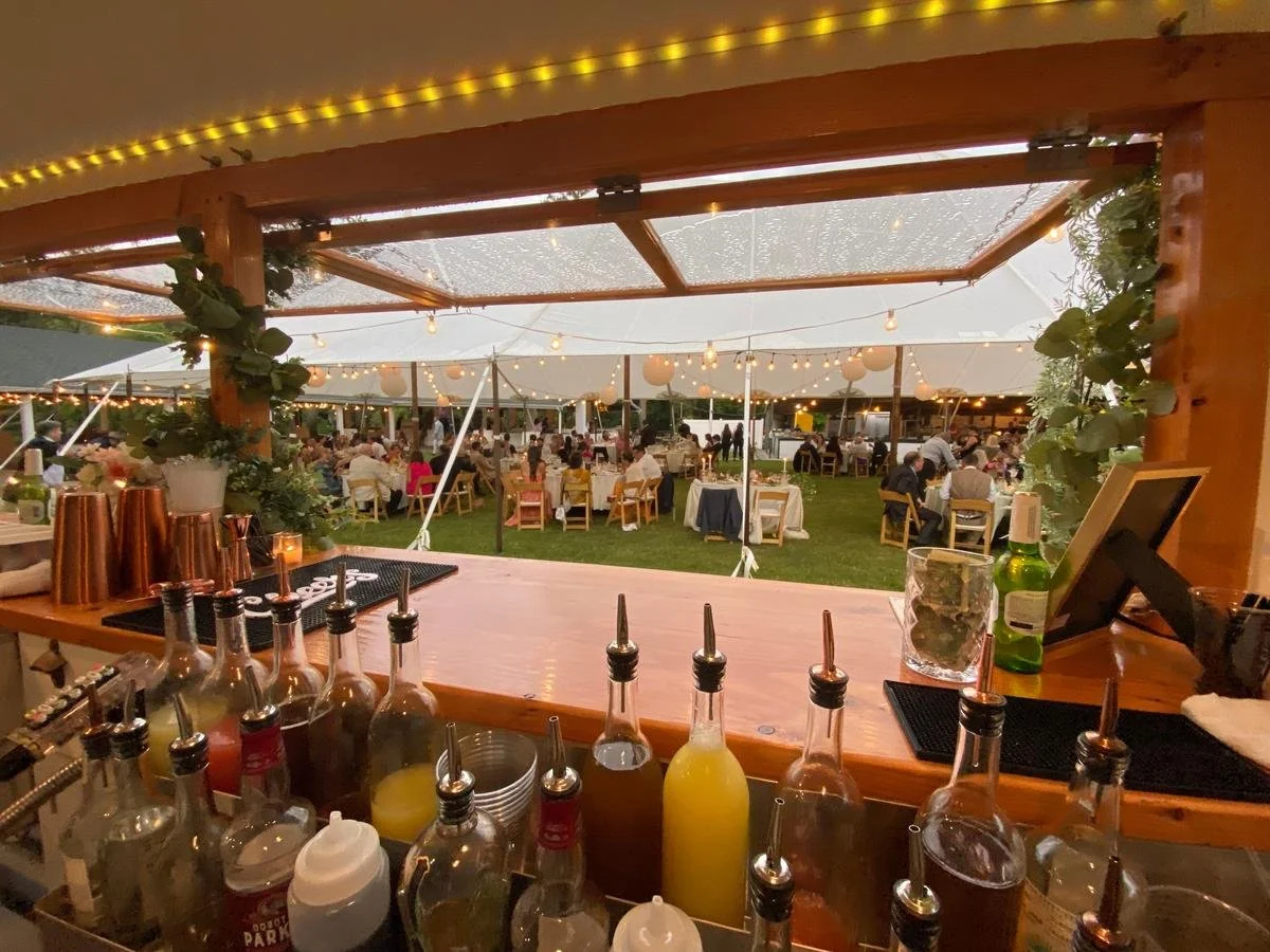 View from a bar into an outdoor wedding reception under a large tent with string lights and guests seated at tables.
