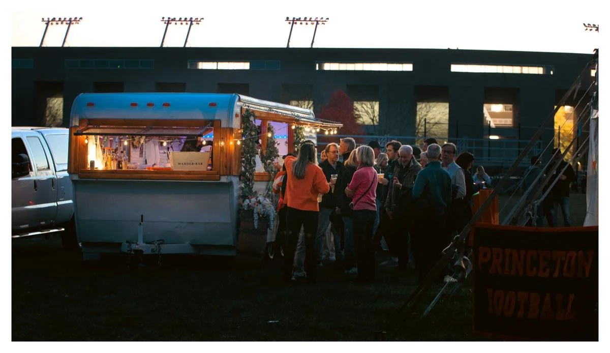 Group of people socializing near a food truck at dusk, with a Princeton football sign in the foreground.