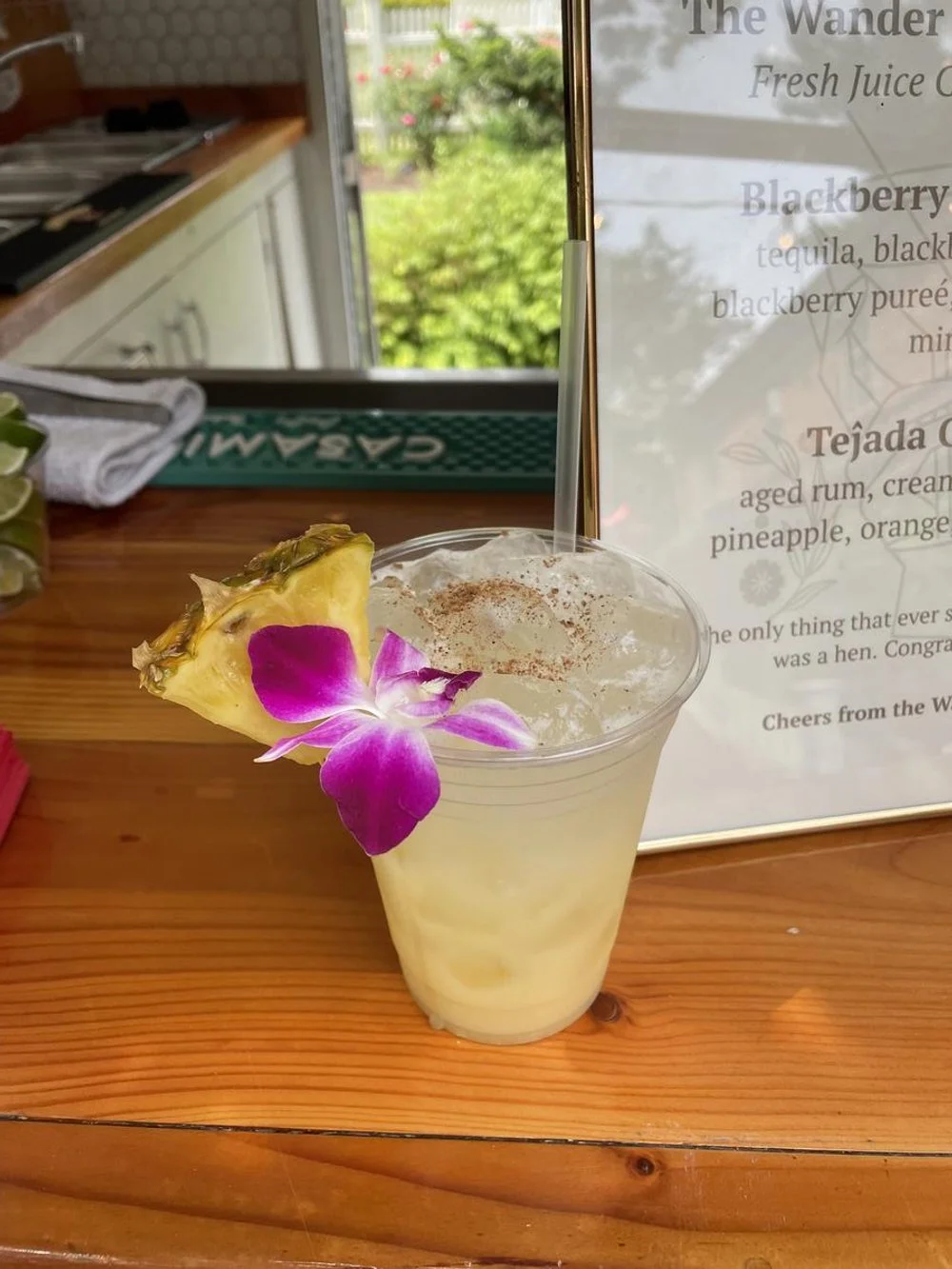 A tropical cocktail garnished with a pineapple slice and a purple orchid flower, sitting on a wooden bar counter.