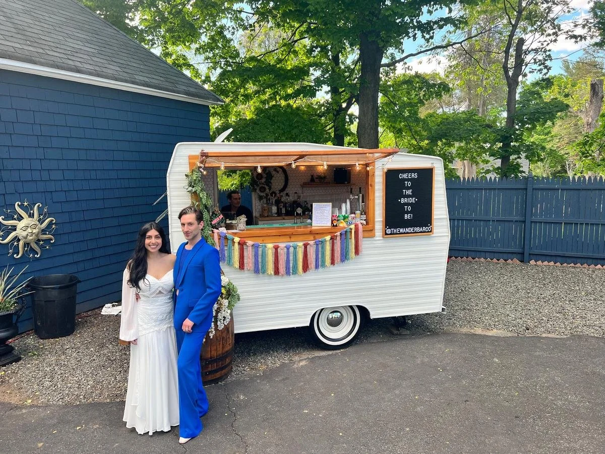A couple in wedding attire standing in front of a small white vintage camper decorated with a colorful tassel garland and a chalkboard sign that reads 'Cheers to the bride to be!'. The wedding couple is smiling, with the woman in a white dress and th