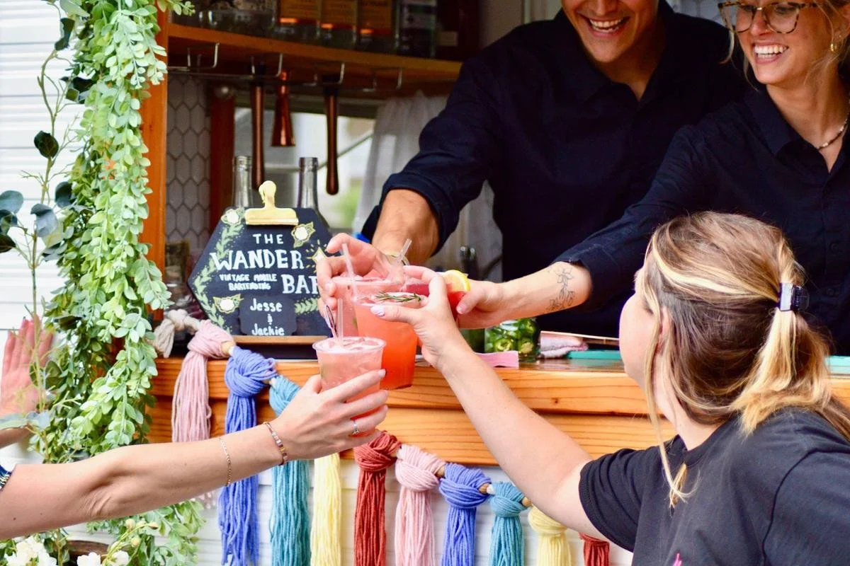 People smiling and toasting with pink drinks at a bar or outdoor stand decorated with colorful yarn knots and greenery.