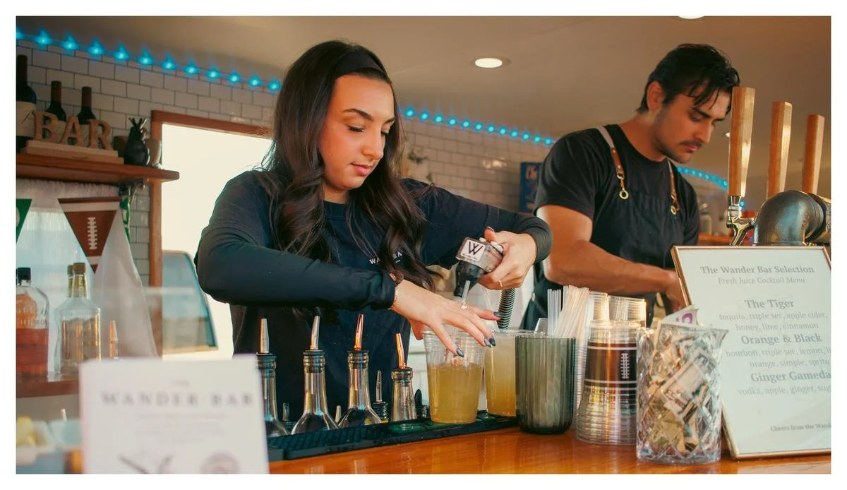 Two bartenders preparing drinks behind a bar with liquor bottles, straws, and a menu.