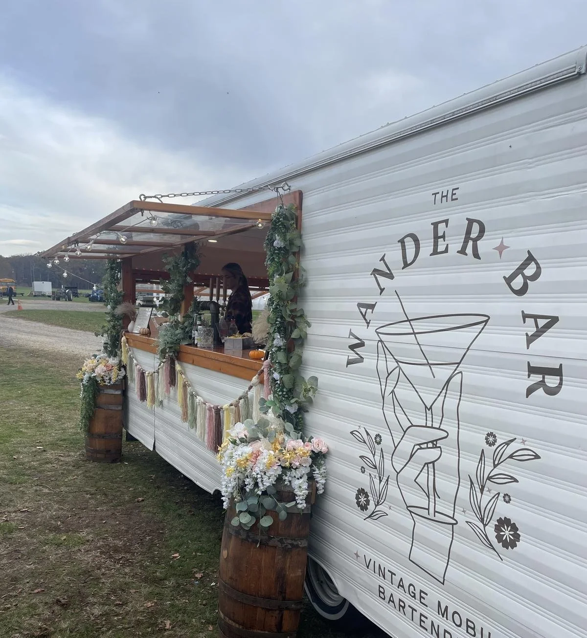 A vintage mobile bar trailer decorated with flowers and greenery, with a serving window and a person behind the counter, set outdoors under cloudy skies.