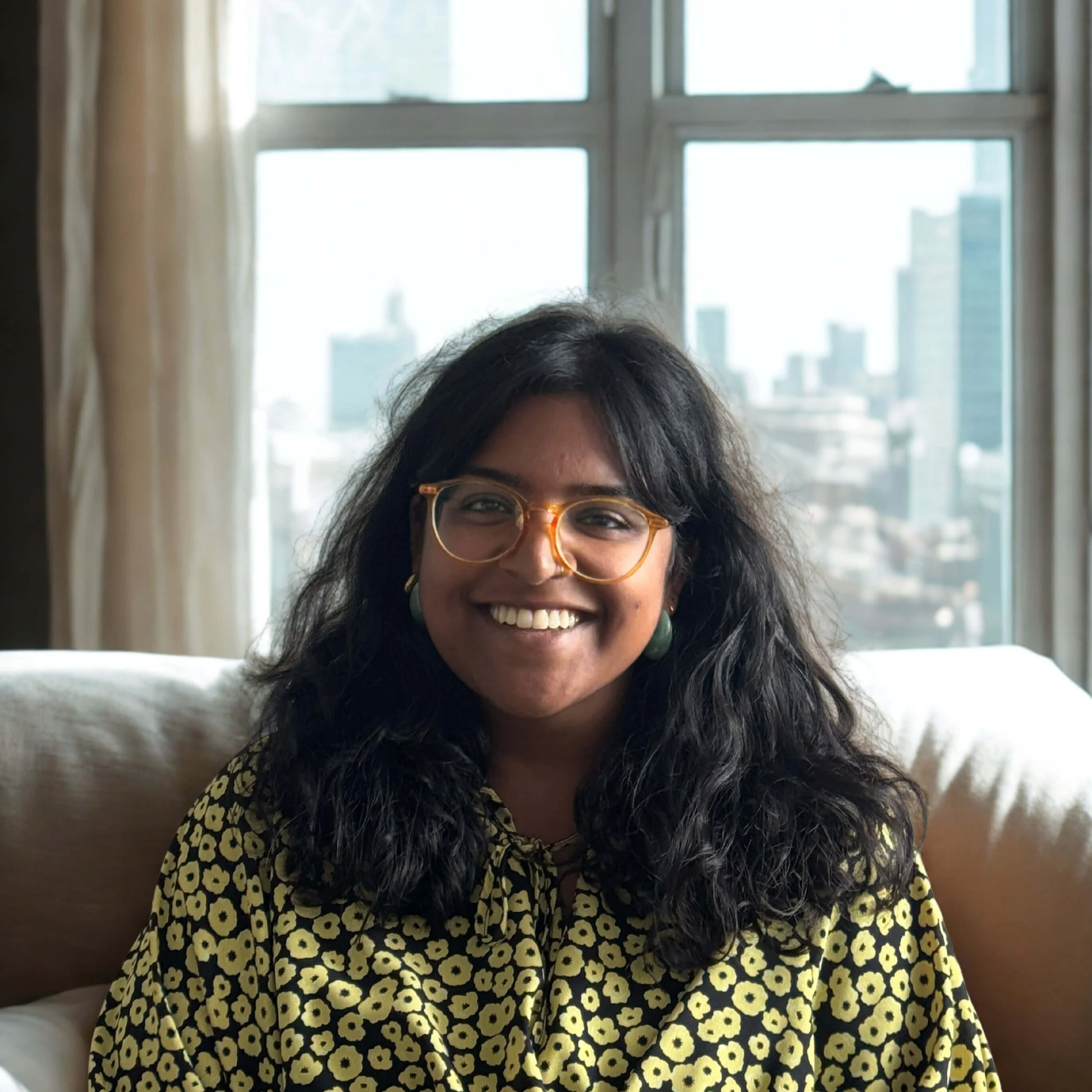 A woman with dark, curly hair, wearing orange glasses and green earrings, smiling while sitting on a beige couch in front of a window with a city skyline in the background.