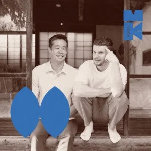 Two men in a traditional Japanese room with shoji screens, one sitting and the other crouching, both smiling, with blue shapes in the foreground and a Japanese logo in the top right corner.