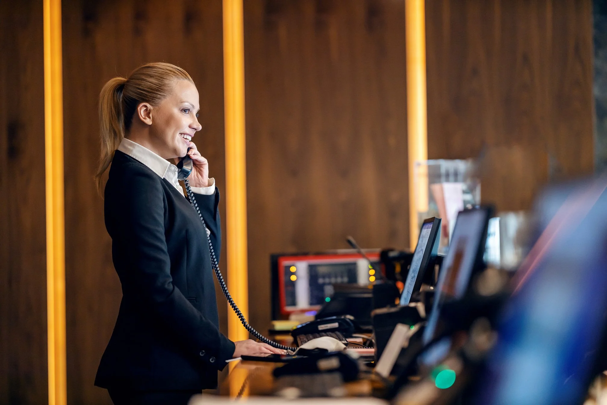 A smiling woman in a hote is talking on the phone at a reception desk with multiple computer screens.
