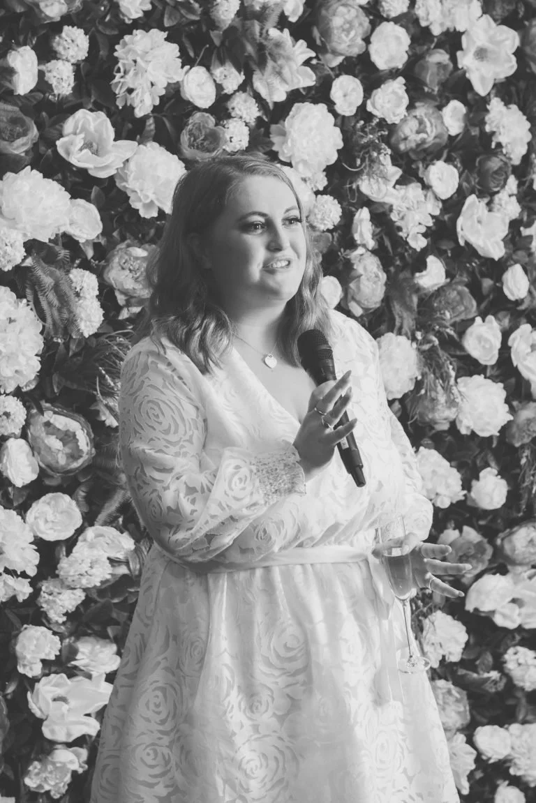 A woman in a floral lace dress holding a microphone and a wine glass, standing in front of a large floral backdrop.