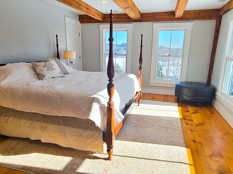 A cozy bedroom with a wooden four-poster bed, white bedding, and decorative pillows. The room has large windows with a scenic view of trees and mountains outside. There's a small lamp on a bedside table, a round black ottoman in the corner, and a bei