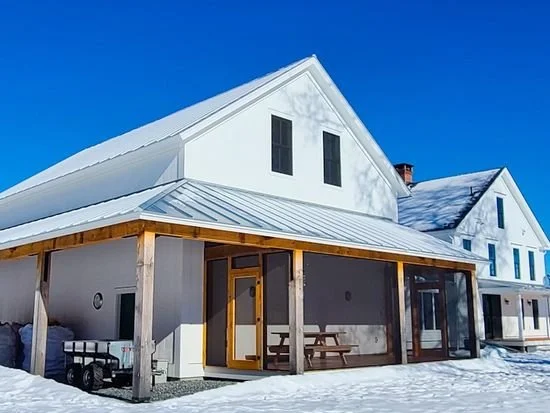 A white house with a metal roof and a covered porch in a snowy landscape, under a clear blue sky.