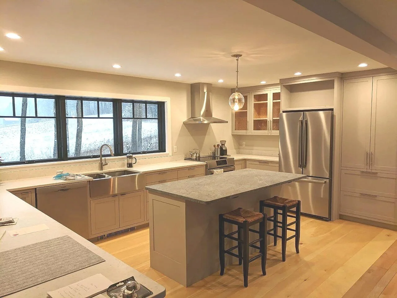Modern kitchen with large window, gray cabinets, stainless steel refrigerator, island with two stools, and a view of a snowy outdoor scene.