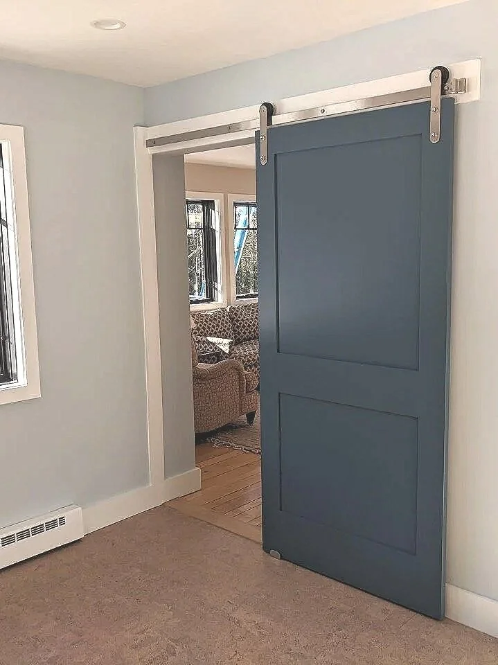 Blue sliding barn door on a white wall, partially open to reveal a living room with large windows and patterned sofas.