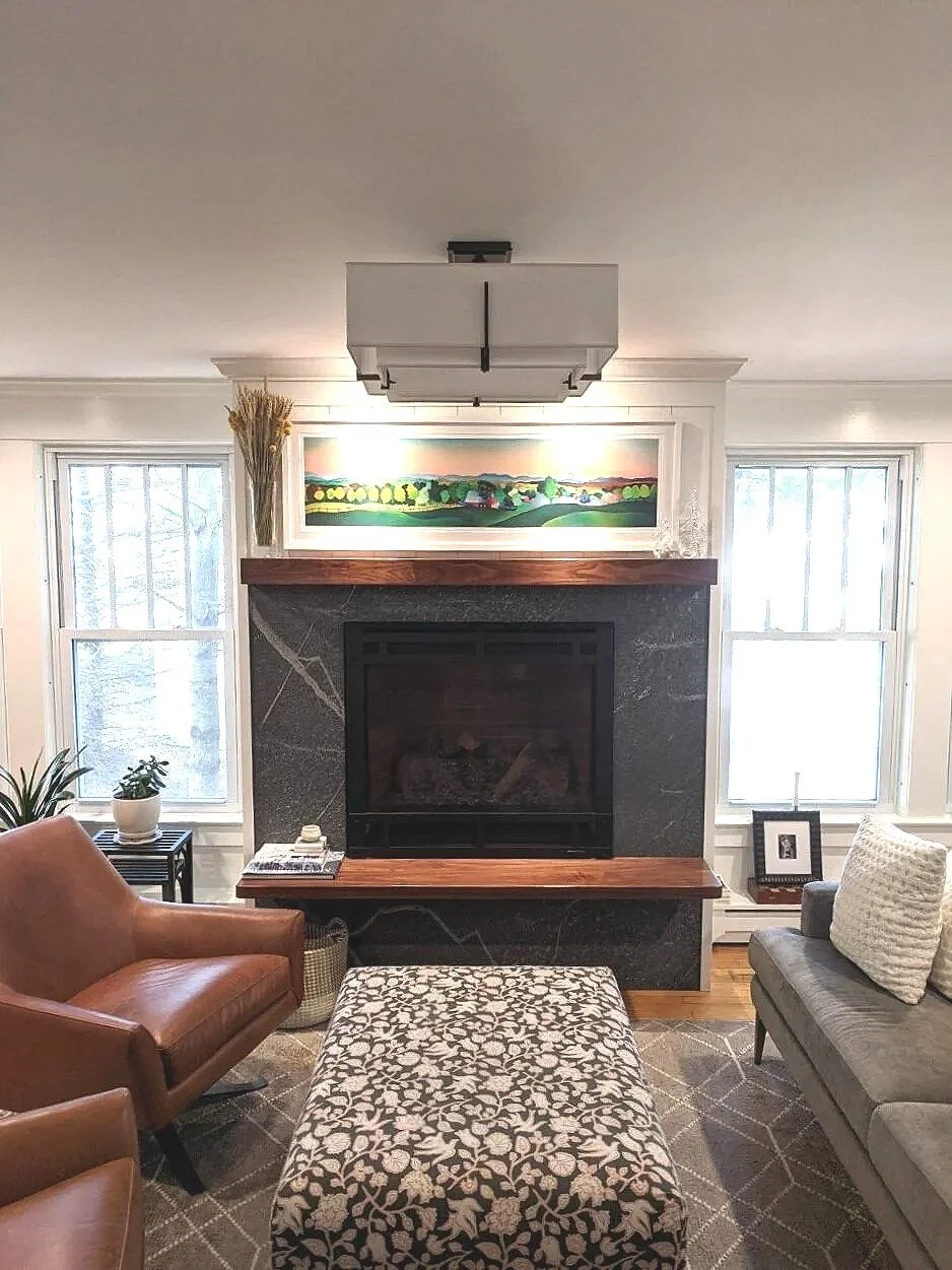 Living room with a fireplace, two large windows, a brown leather chair, a gray sofa with pillow, a black side table with plants, and a patterned ottoman in front of the fireplace, which has a colorful landscape painting above it.