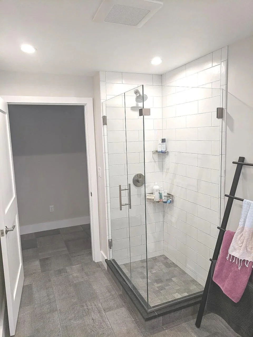 A modern bathroom with a glass shower enclosure, white tiled walls, gray tile flooring, and a black towel rack with towels.