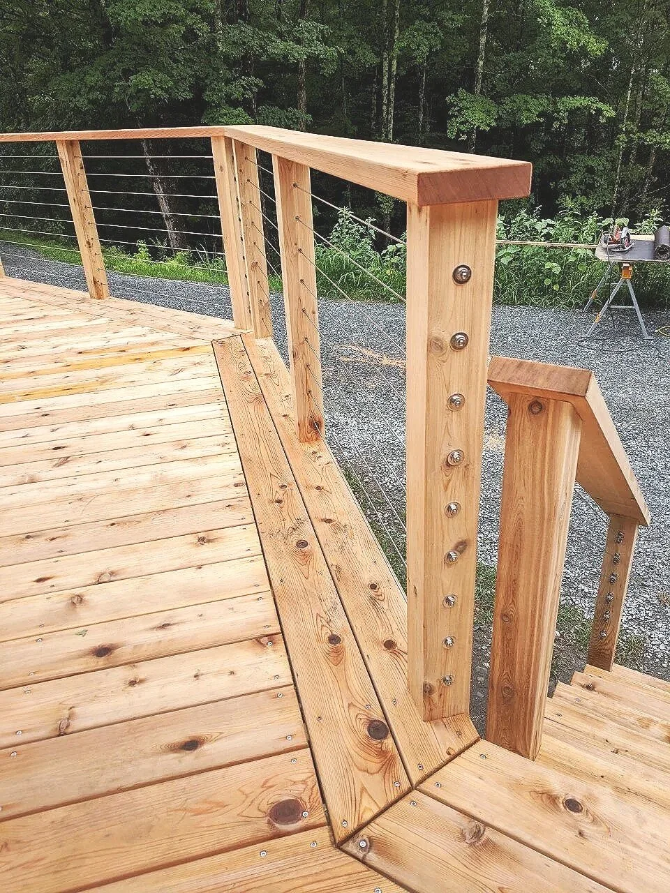 Close-up view of a wooden deck with a metal cable railing, with stairs leading down and a forest or wooded area in the background.