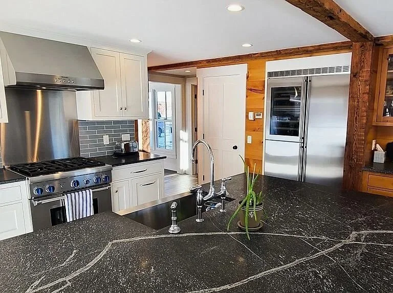 Modern kitchen with white cabinetry, black countertops, stainless steel appliances including a large refrigerator and stove, gray tile backsplash, and a black island with a sink and a small plant.