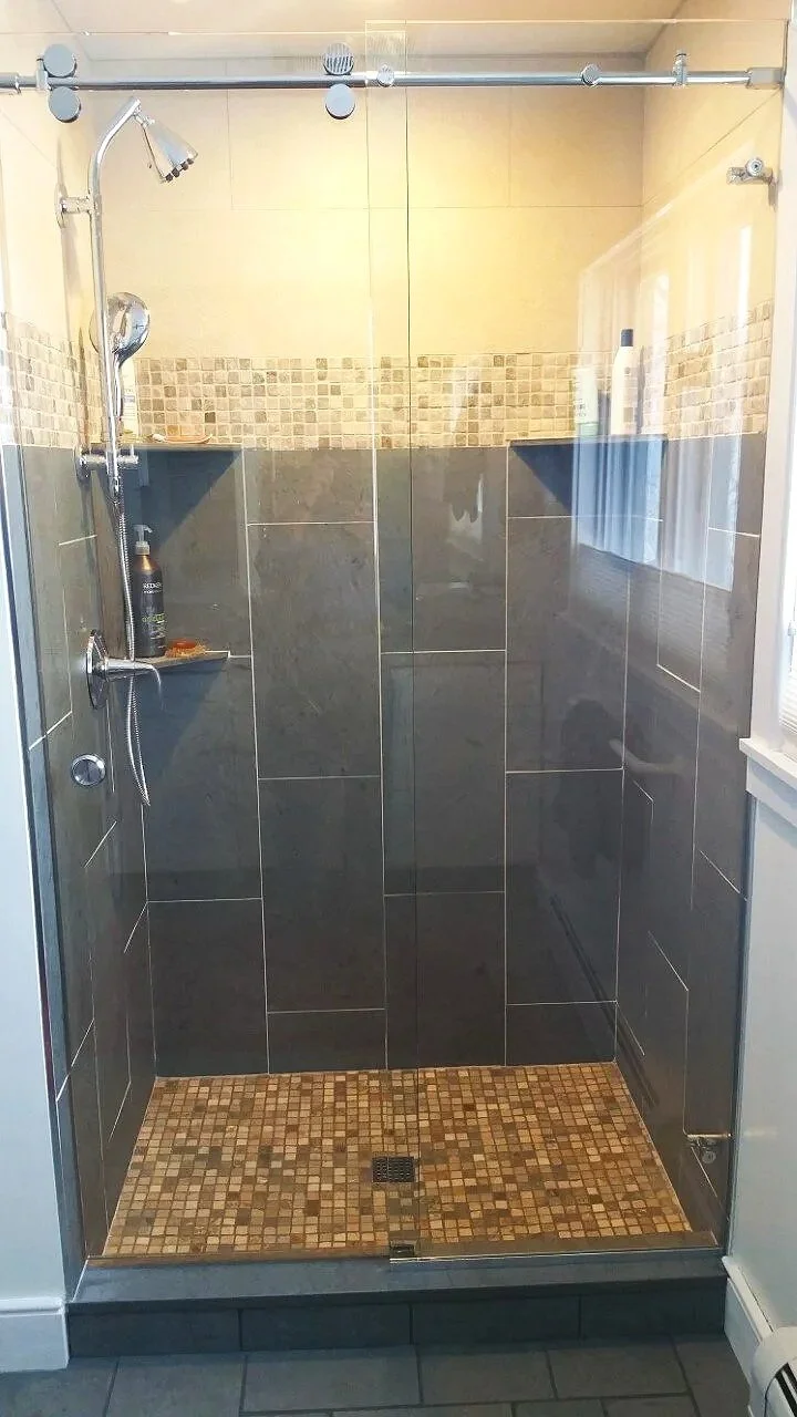 A walk-in shower with dark gray tiles and mosaic floor tiles, two corner shelves holding bottles of shampoo and body wash, and a sliding glass door with a metal frame.