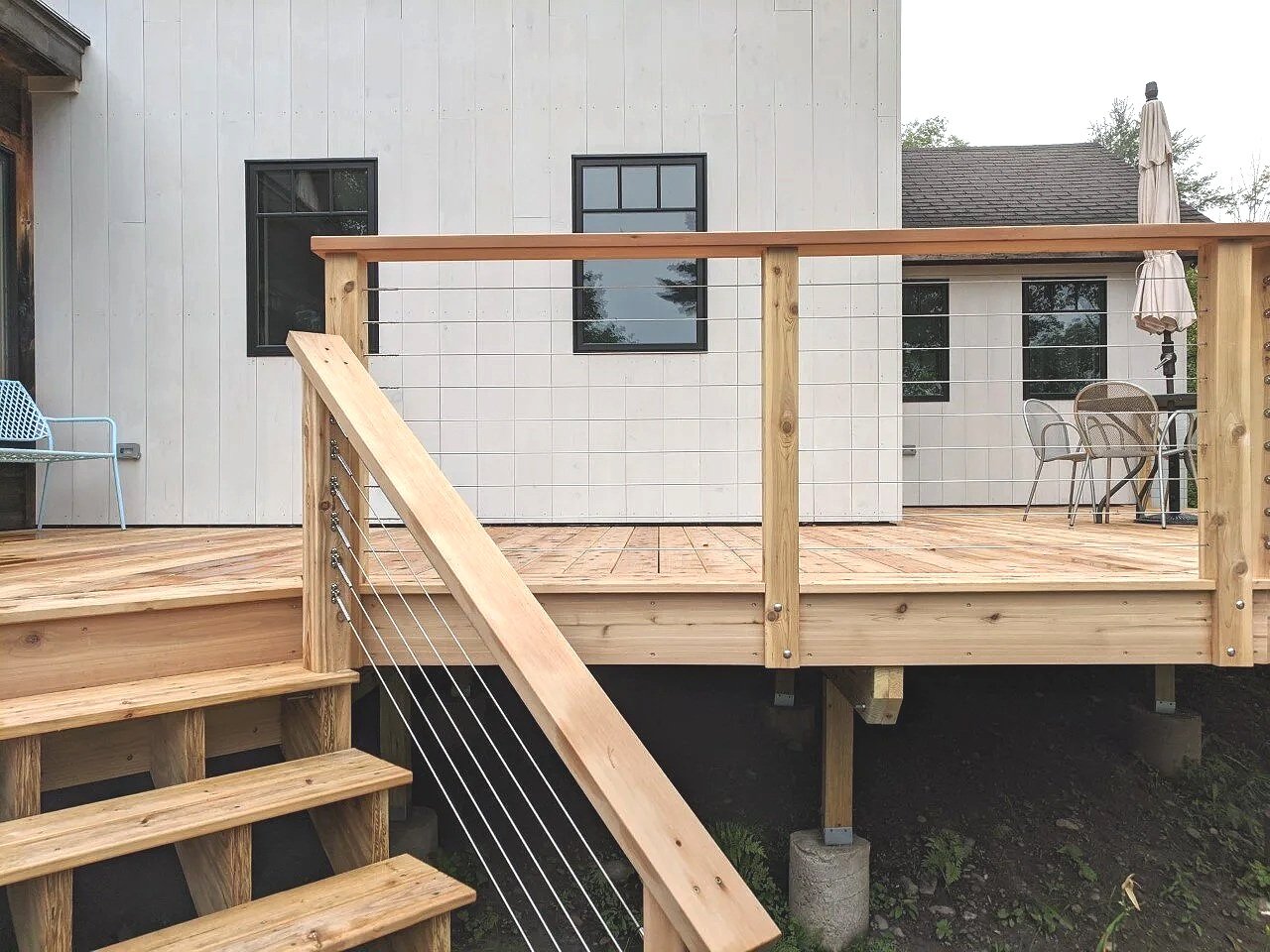 Wooden outdoor deck with stairs, railing, patio furniture, and an umbrella adjacent to a white house with black windows.