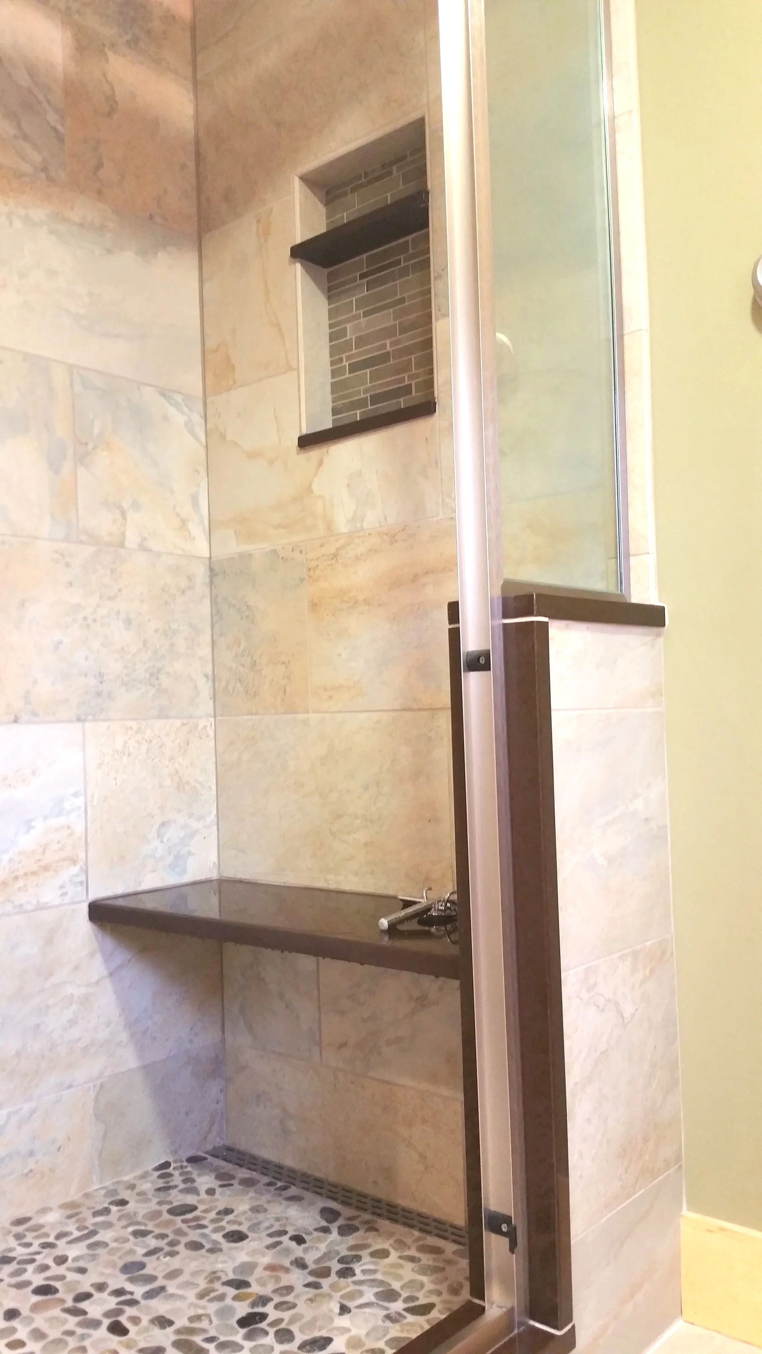 Inside a shower with beige stone tiles, a small built-in niche with dark tile, a dark brown shelf, and a pebble tiled shower floor.