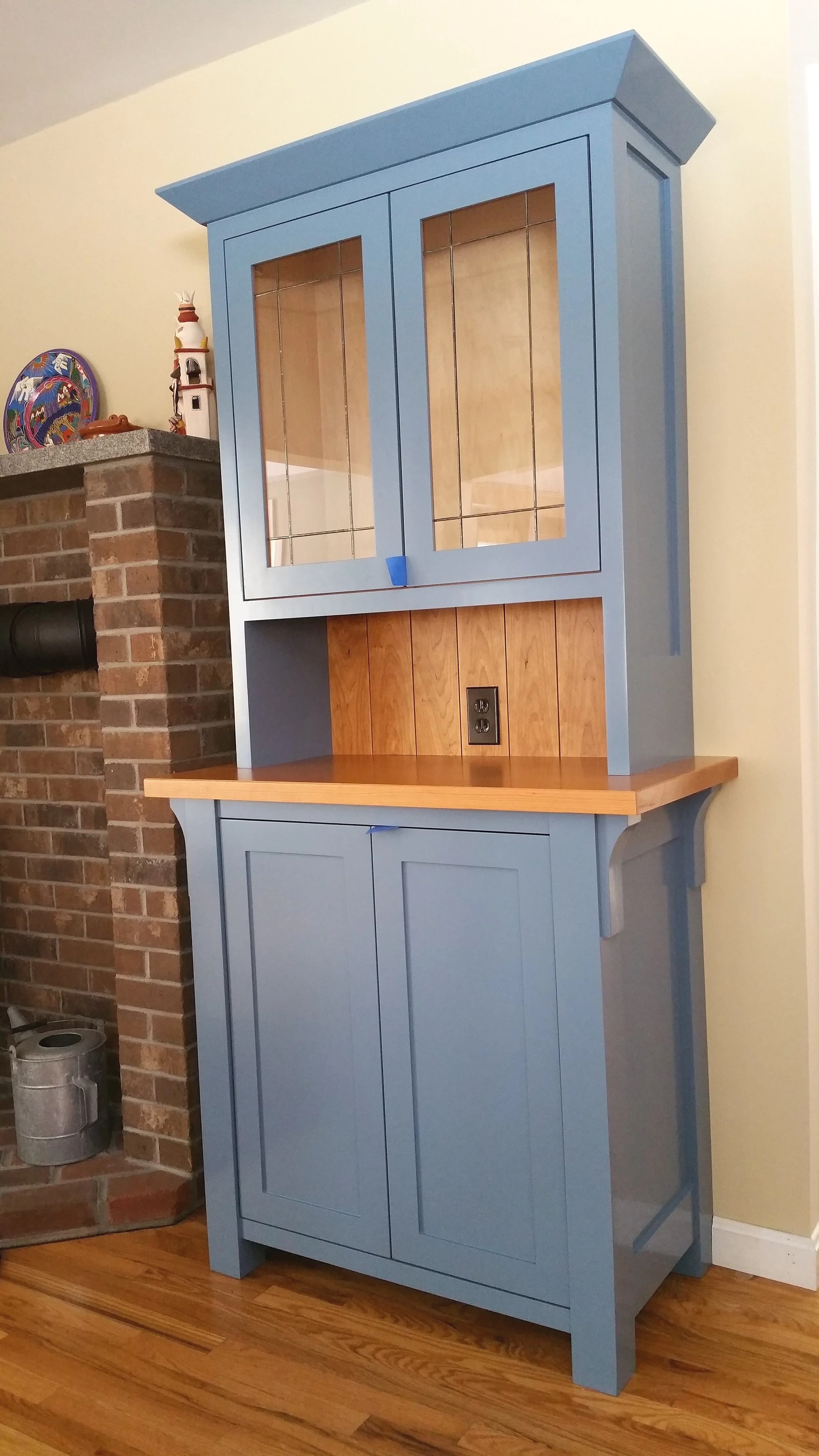 A tall, blue wooden cabinet with glass doors on the upper section and solid doors on the lower section, with a wooden countertop and backsplash, in a room with hardwood flooring and a brick wall.