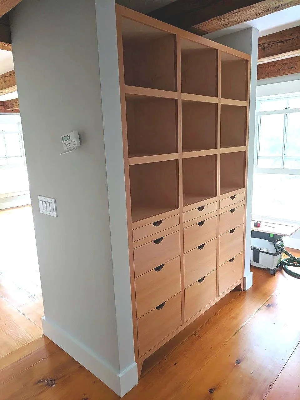A wooden open shelving unit with multiple compartments and drawers, positioned next to a white wall with a thermostat and light switches, inside a room with wooden floors and large windows.