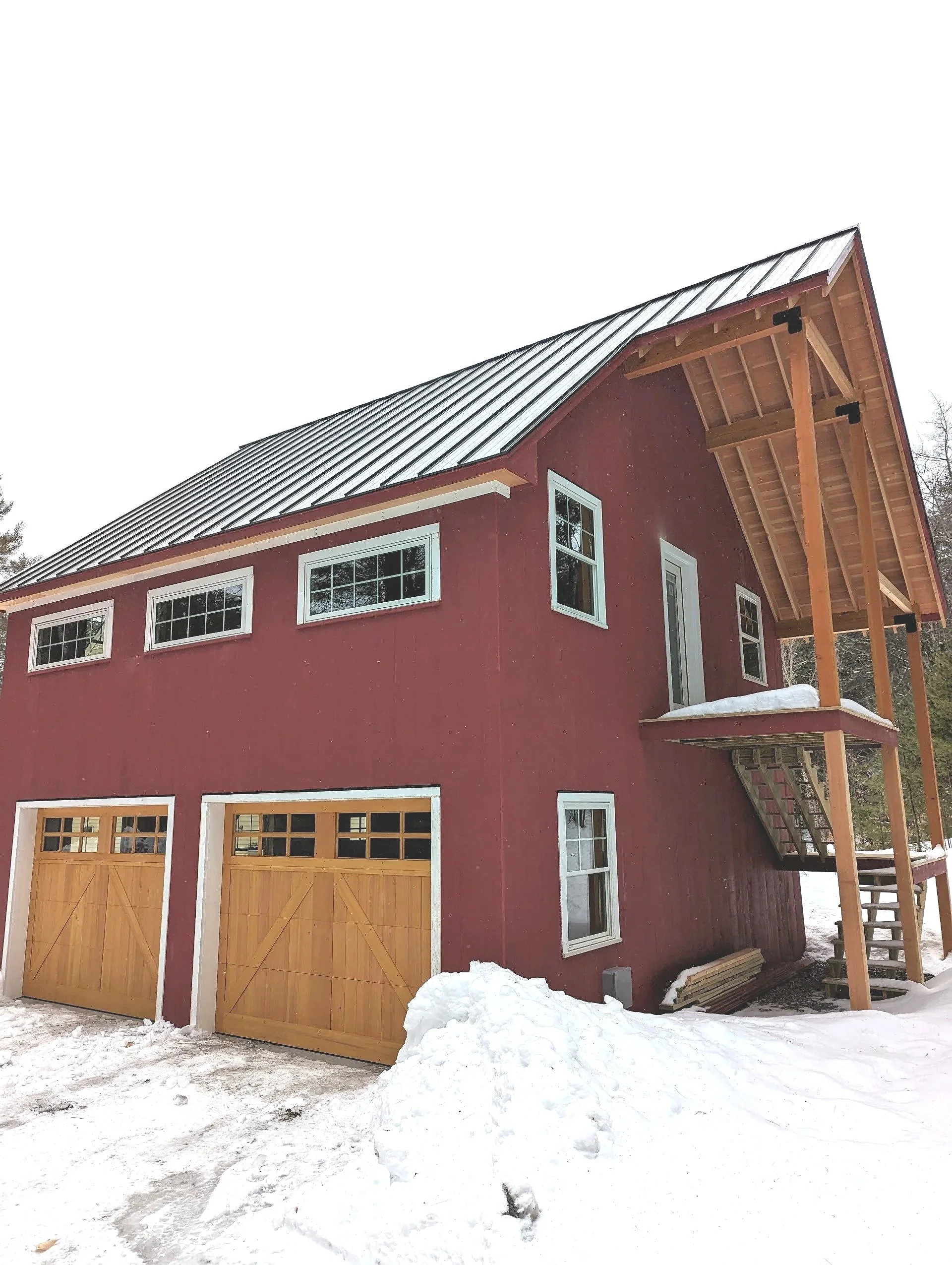 A two-story house painted red with white trim and wooden garage doors, snow on the ground and on the house's roof, with a wooden staircase leading to a side door and a snow-covered deck.