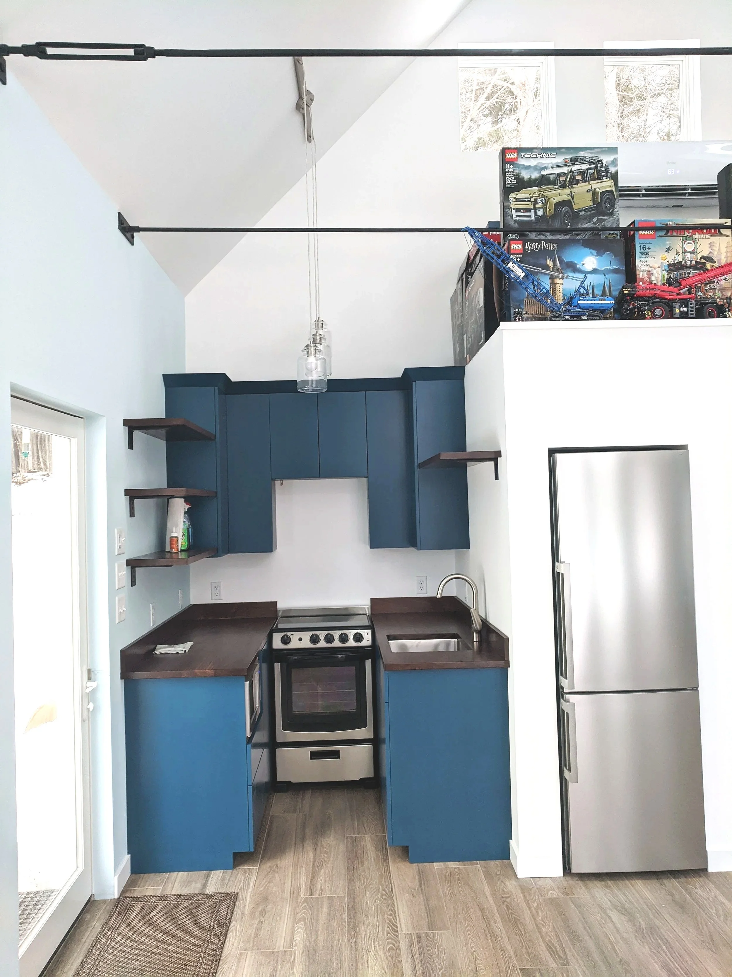 Small kitchen with blue cabinets, dark wood countertops, stainless steel appliances, and open shelves. Overhead, motorized craft project tracks hold Lego construction sets, including a yellow vehicle, blue crane, and red truck, on a loft above the ki