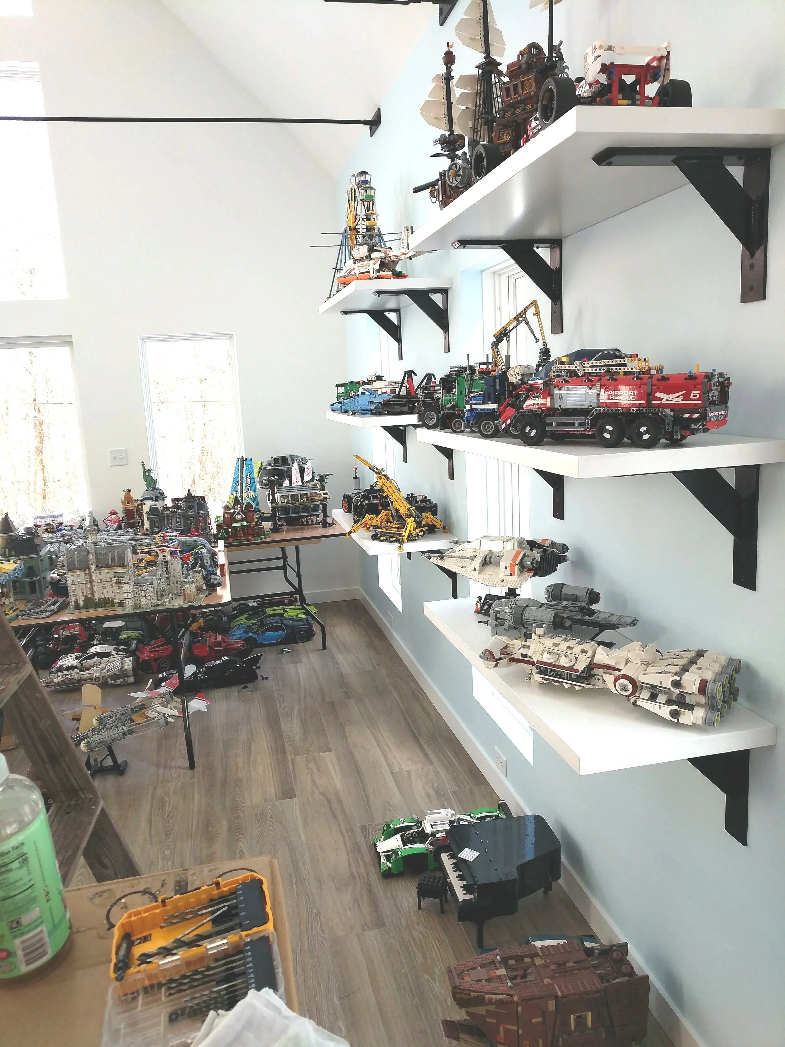 Room with white walls and wooden flooring, displaying several white floating shelves filled with colorful LEGO model ships, cars, and other vehicles. A small black piano and a wooden box are on the floor, along with a table with additional LEGO model