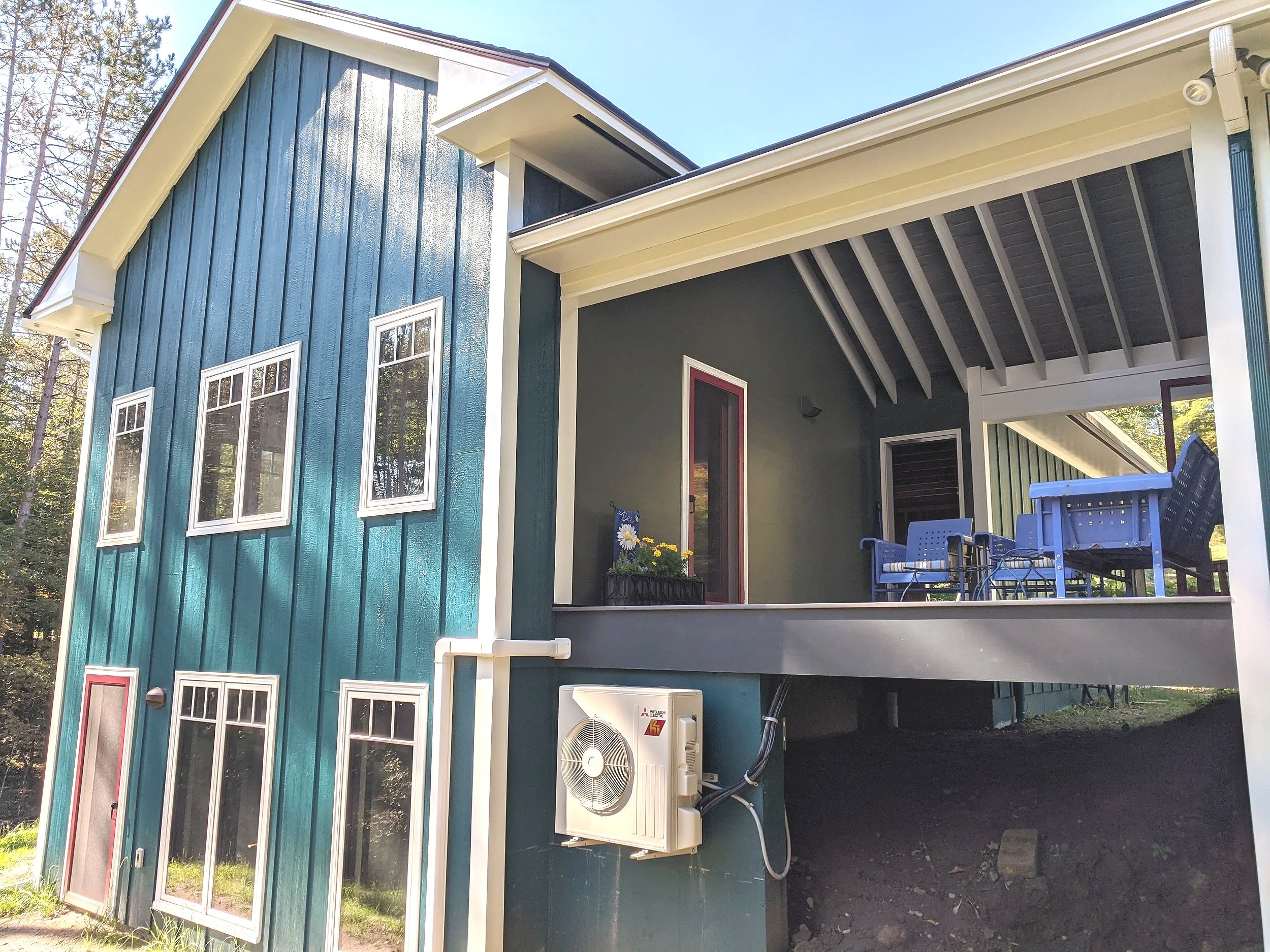 Side view of a two-story house with teal siding, white trim, and a covered porch with blue outdoor furniture. The house has six windows, three on the lower level and three on the upper level. An air conditioning unit is mounted on the exterior wall n