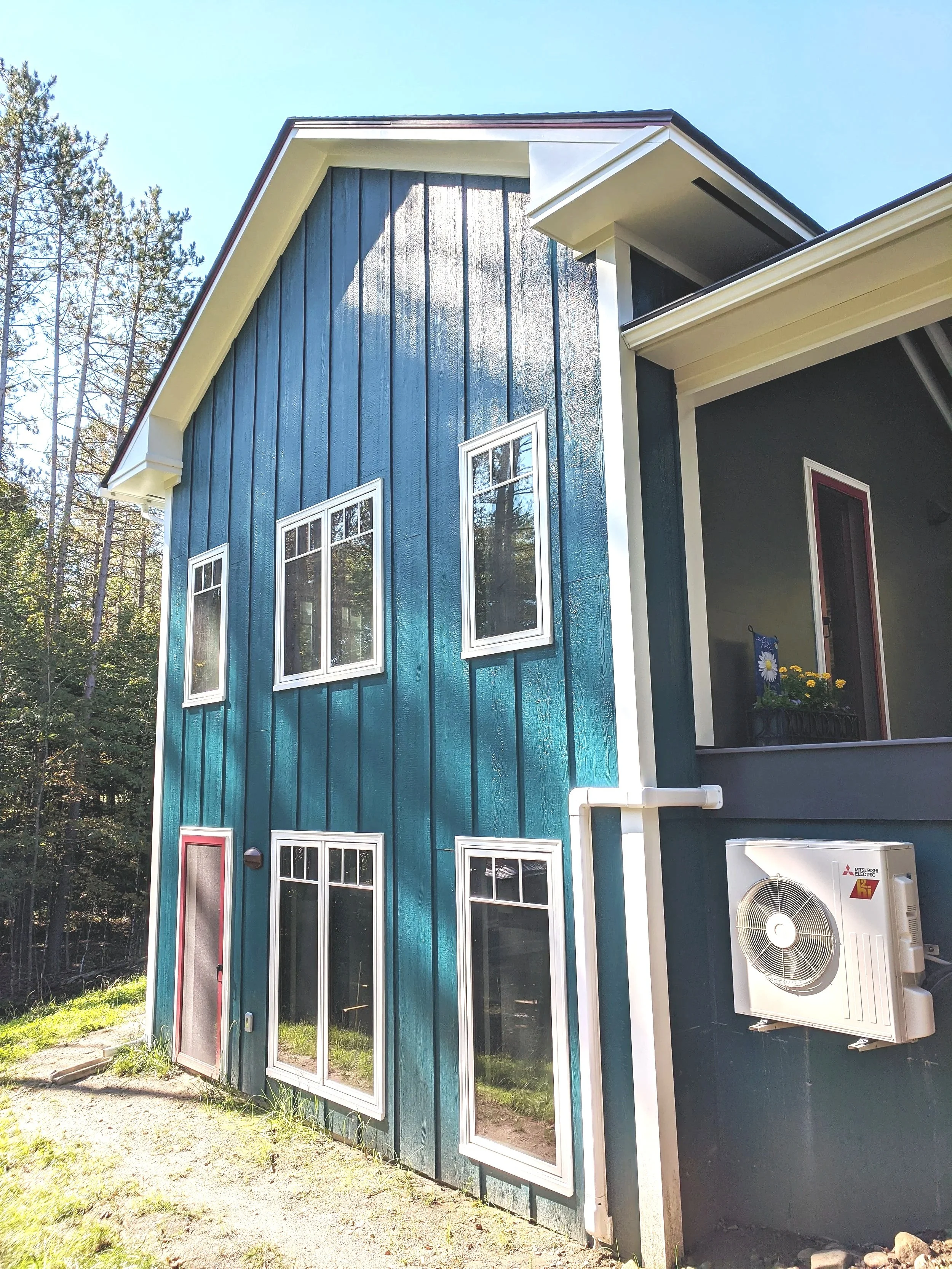 Front exterior view of a modern two-story house with blue vertical siding, multiple white-framed windows, and a small porch with potted flowers. Air conditioning unit is mounted on the side.