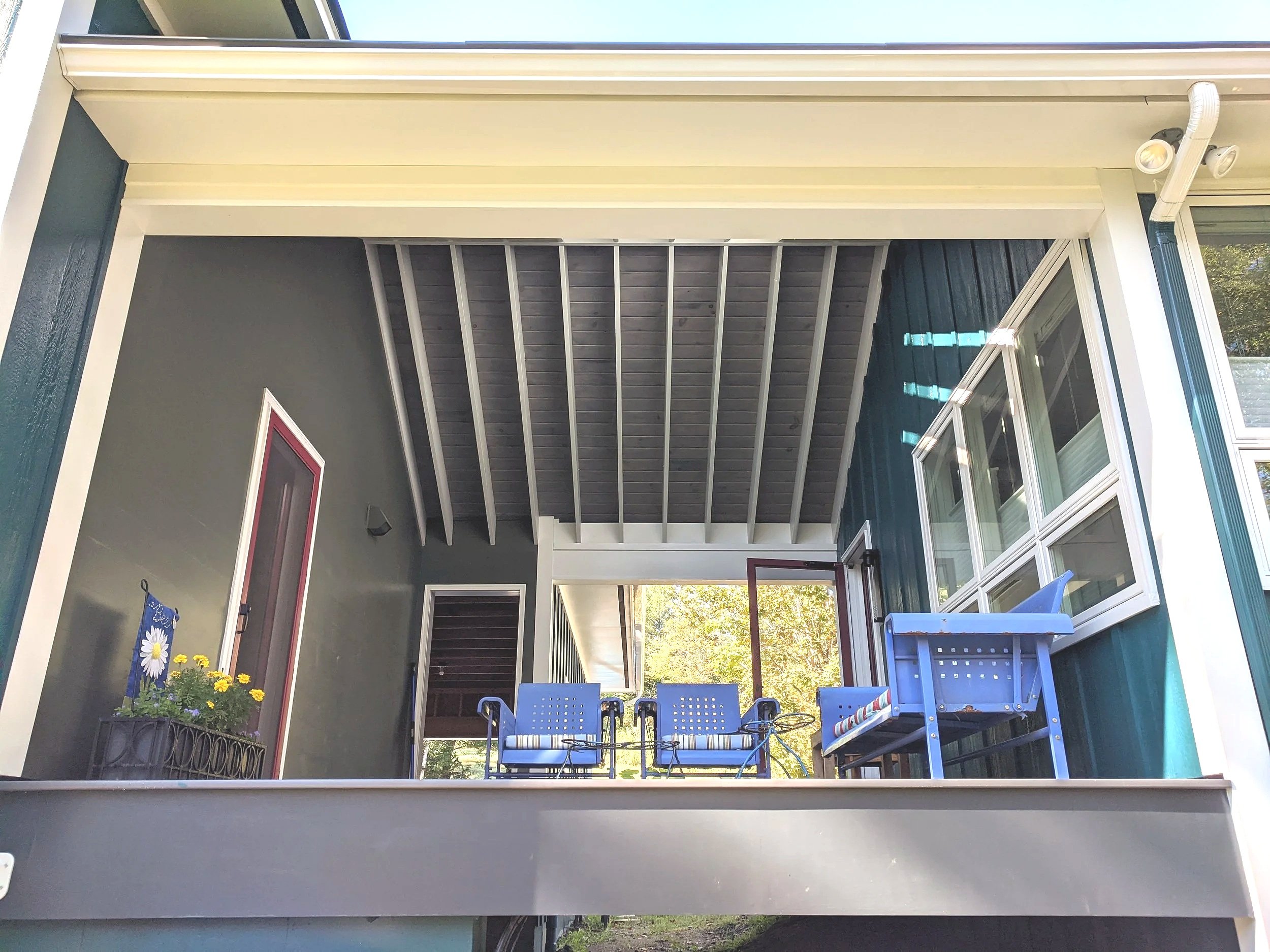 View of a second-story balcony with outdoor furniture, including blue chairs and a small blue table, with windows and plants visible.